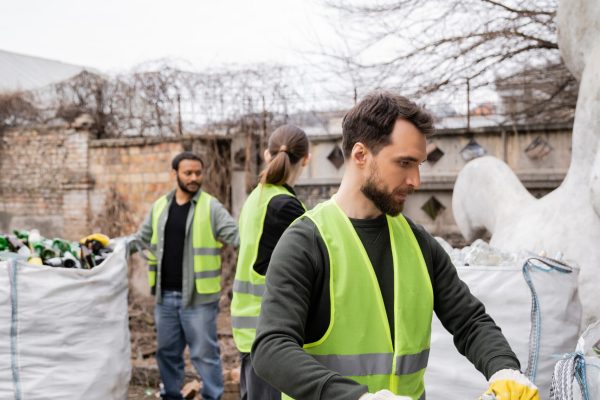 Bearded worker in safety vest and gloves holding sack with glass trash near blurred multiethnic colleagues in outdoors waste disposal station, garbage sorting and recycling concept