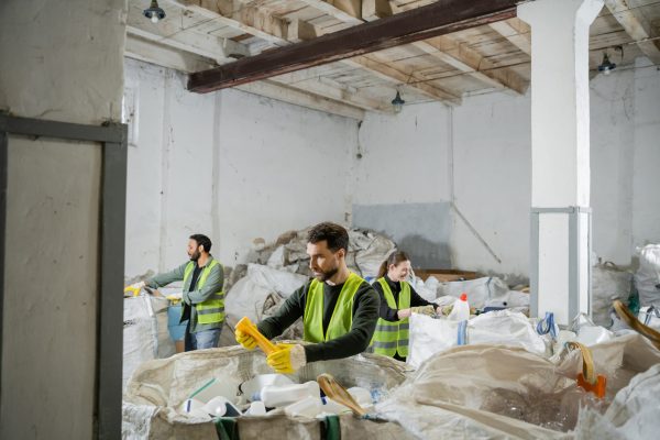 Male worker in protective vest and gloves holding plastic trash while standing near sacks and