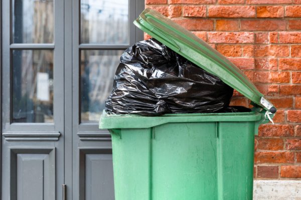 Opened big green plastic trash can with full trash bags. Disposal of waste. Overcrowded garbage can over brick wall and door.