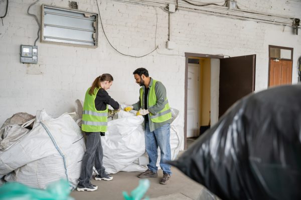 Side view of young worker in high visibility vest and gloves standing near sacks and indian colleague while working in waste disposal station, garbage sorting and recycling concept